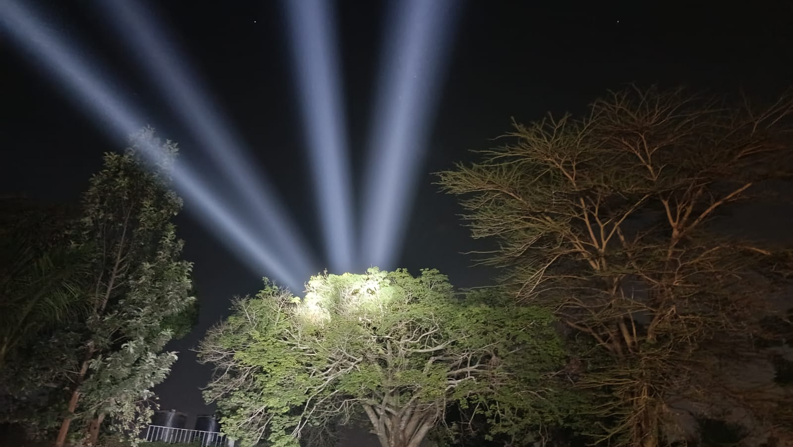 Searchlight beams projected above the treeline at an outdoor evening event in Kenya